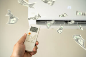 A person's hand holding a remote in front of an air conditioner, adjusting the temperature while money floats around - highlighting air conditioning costs and indicating Waywest Mechanical has some tips to help reduce them.